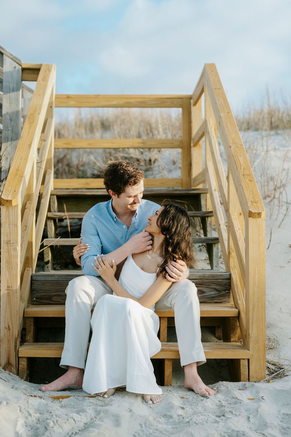 Couple sitting closely on wooden beach stairs, man in blue shirt embracing woman in white dress, sandy dunes background.