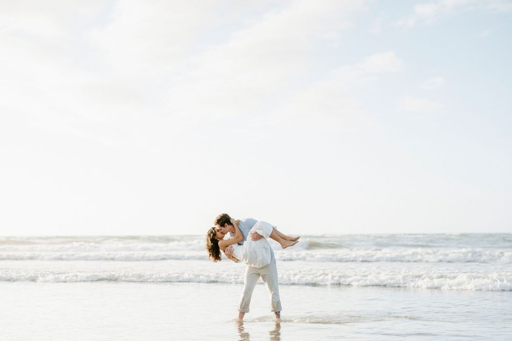 Romantic couple sharing a kiss as man lifts woman at the ocean shoreline during golden hour.