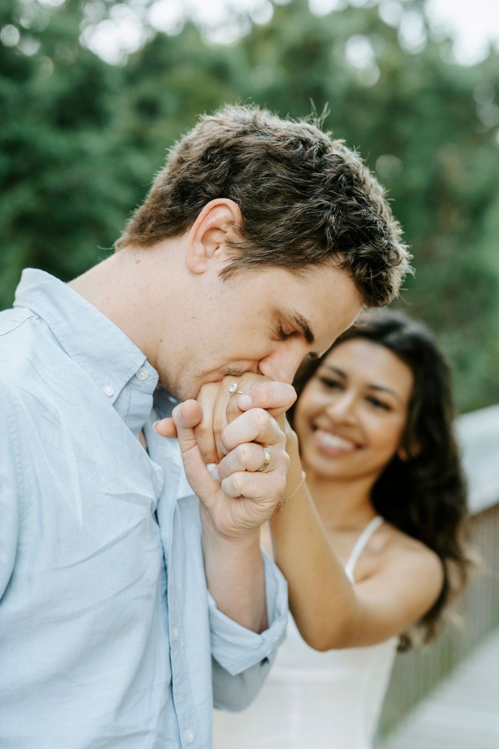 Man in blue shirt tenderly kissing woman's hand while she smiles warmly in outdoor setting.