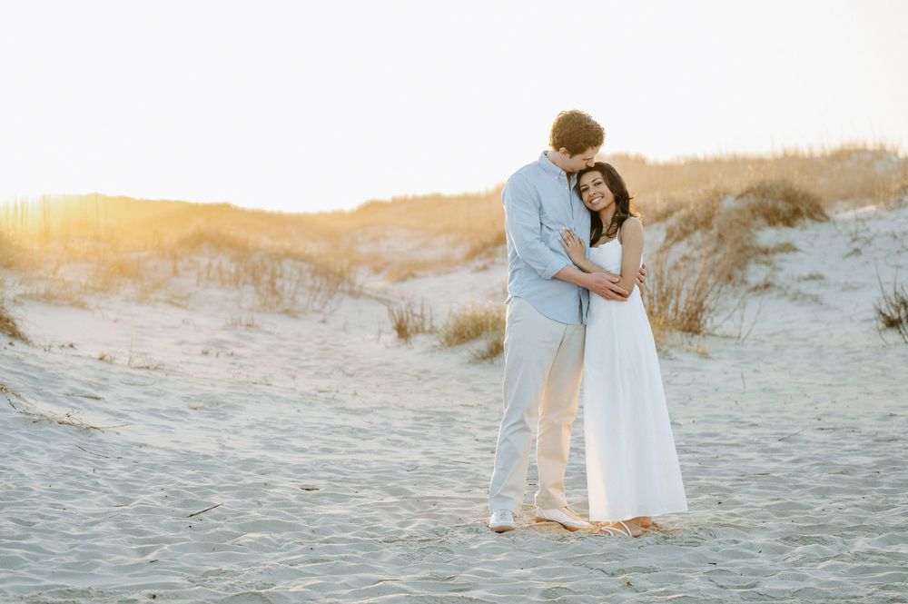 Couple embracing on sandy beach dunes at golden sunset, woman in white dress, man in light blue shirt.