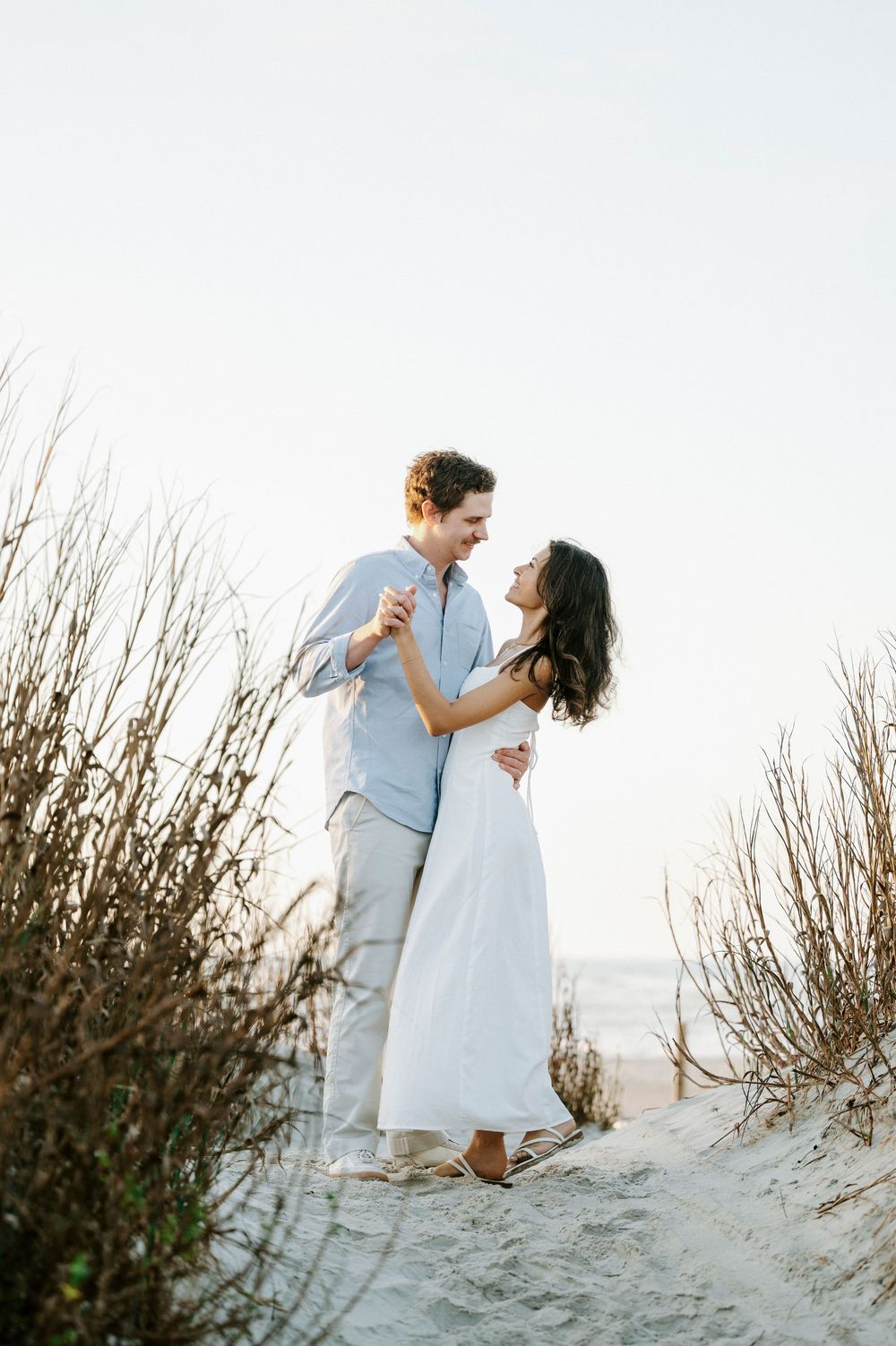 Couple dancing romantically on sandy beach dunes at sunset, woman in white dress, man in light blue shirt.