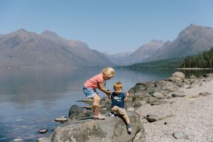 Children exploring rocky shoreline of mountain lake with peaks in background.