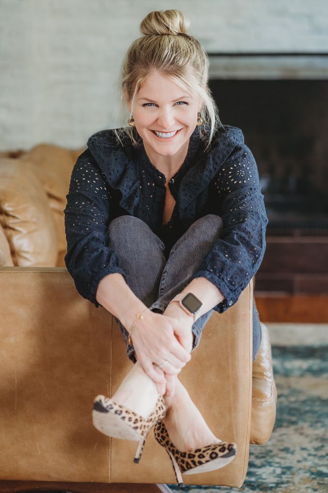 Woman in navy sparkly top and leopard print heels sits casually on tan leather couch with bright smile.
