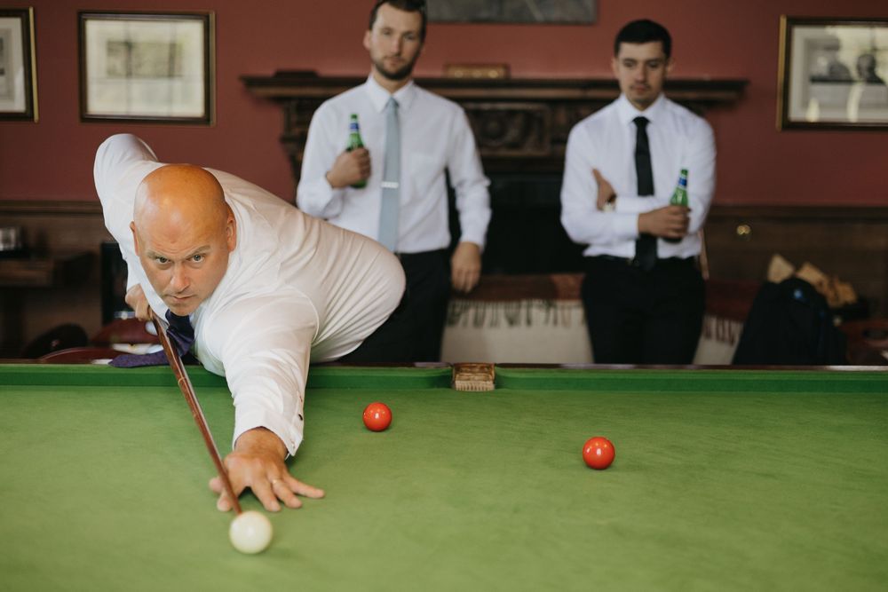 Groomsmen playing pool at a wedding venue with red walls and a classic billiards table.