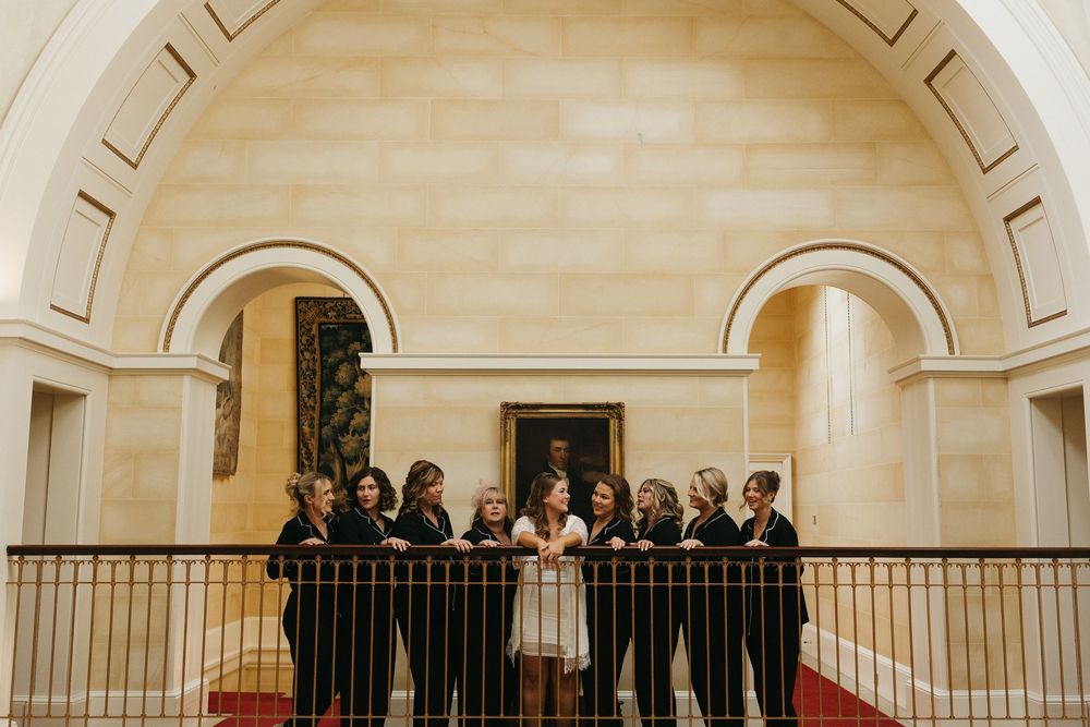 Wedding party stands together on a grand staircase with arched architecture and elegant railings in warm lighting.
