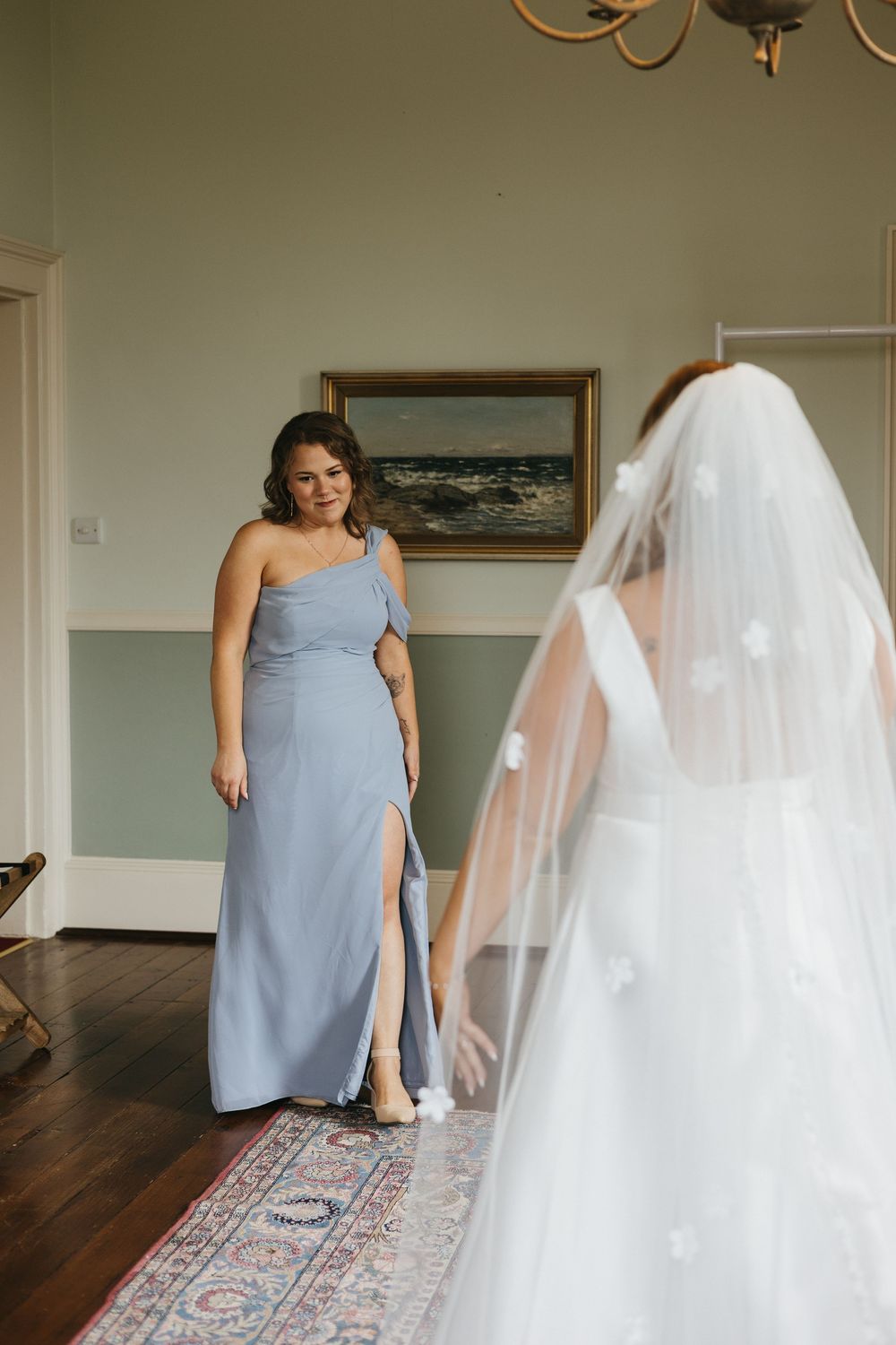 Bridesmaids in powder blue dresses stand on hardwood floor near a white wedding dress and ornate rug.