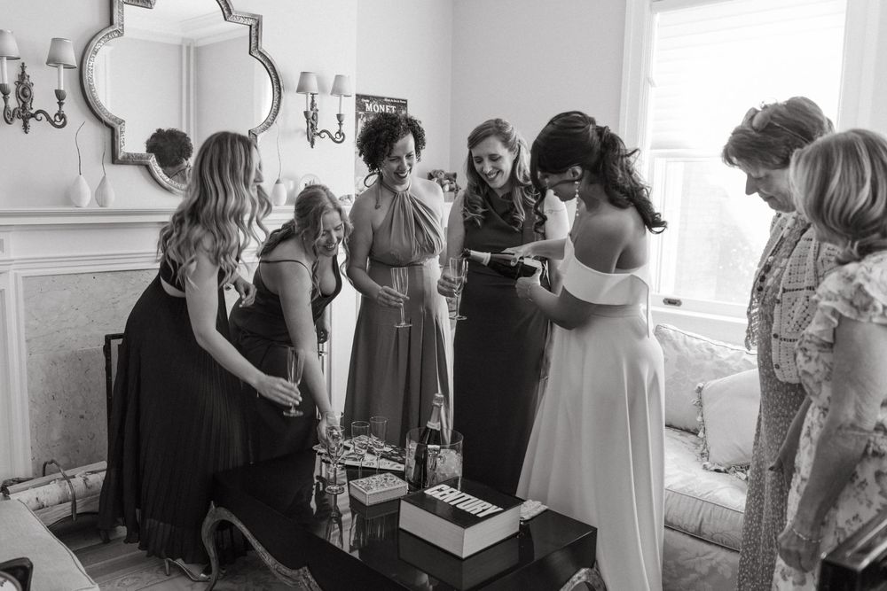 Bridesmaids help adjust a wedding dress in an elegant white room with mirrors and dress racks.
