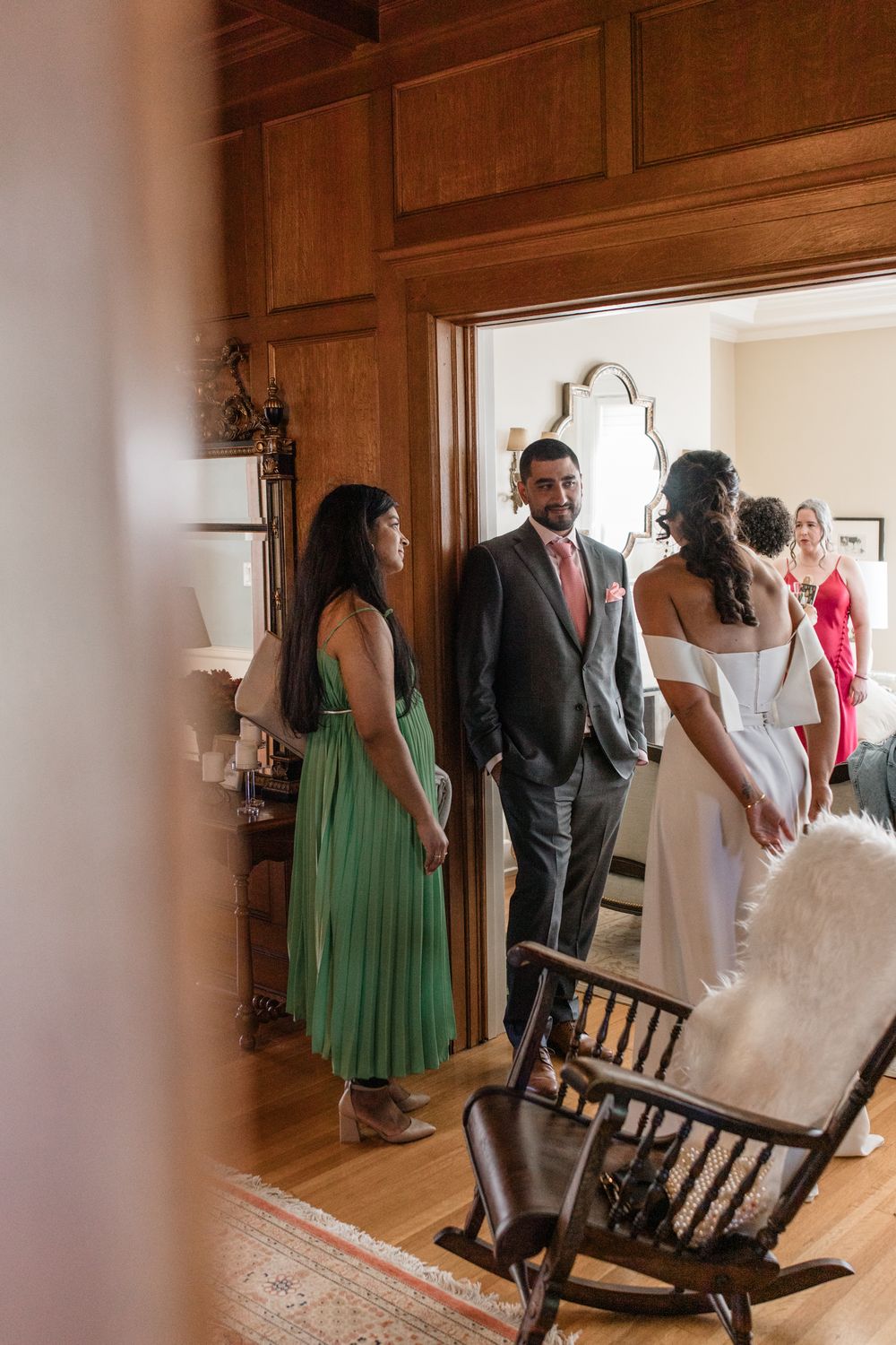 A wedding party in green dresses and formal attire gathers in a wood-paneled room during preparations.