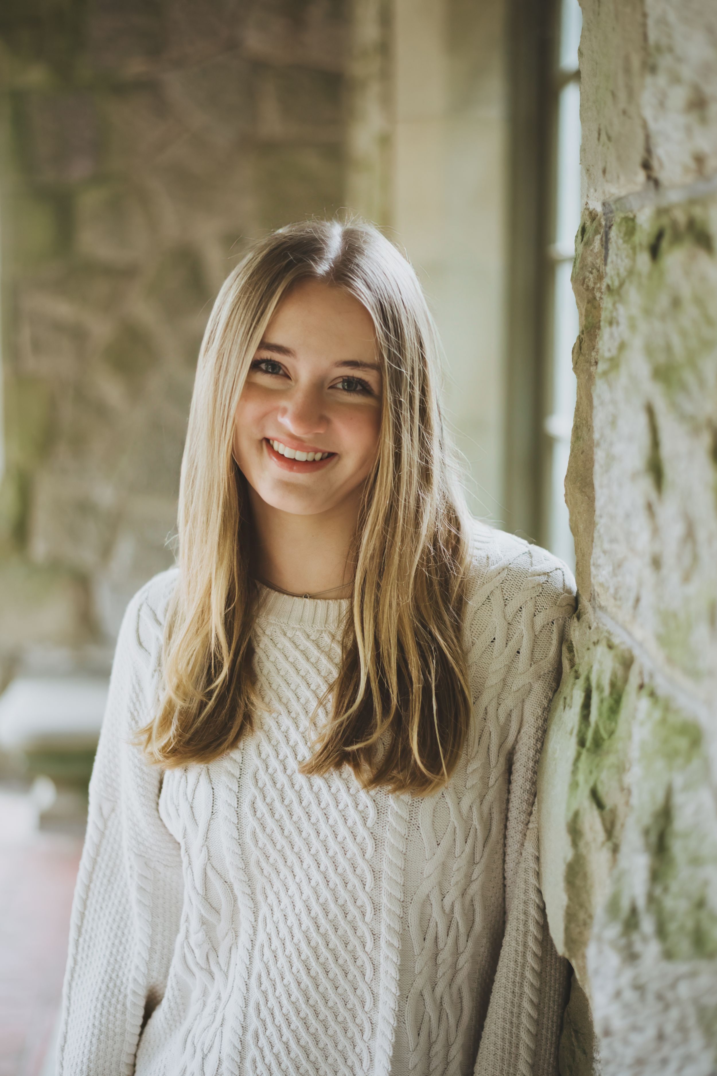 A person wearing a white cable knit sweater smiles while standing against a stone wall in natural lighting.