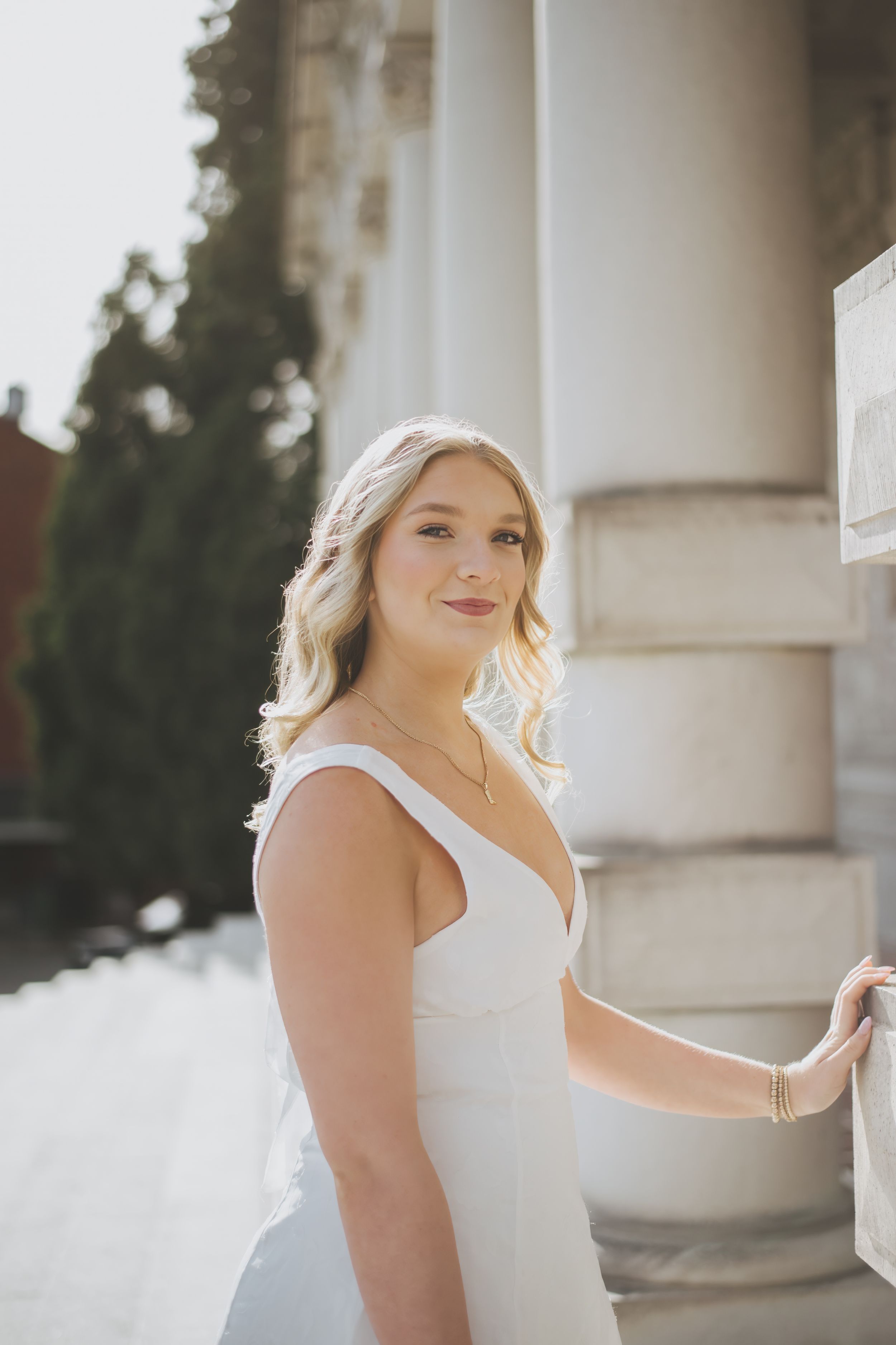 A bride in a white sleeveless wedding dress poses elegantly next to a classic stone column in natural lighting.