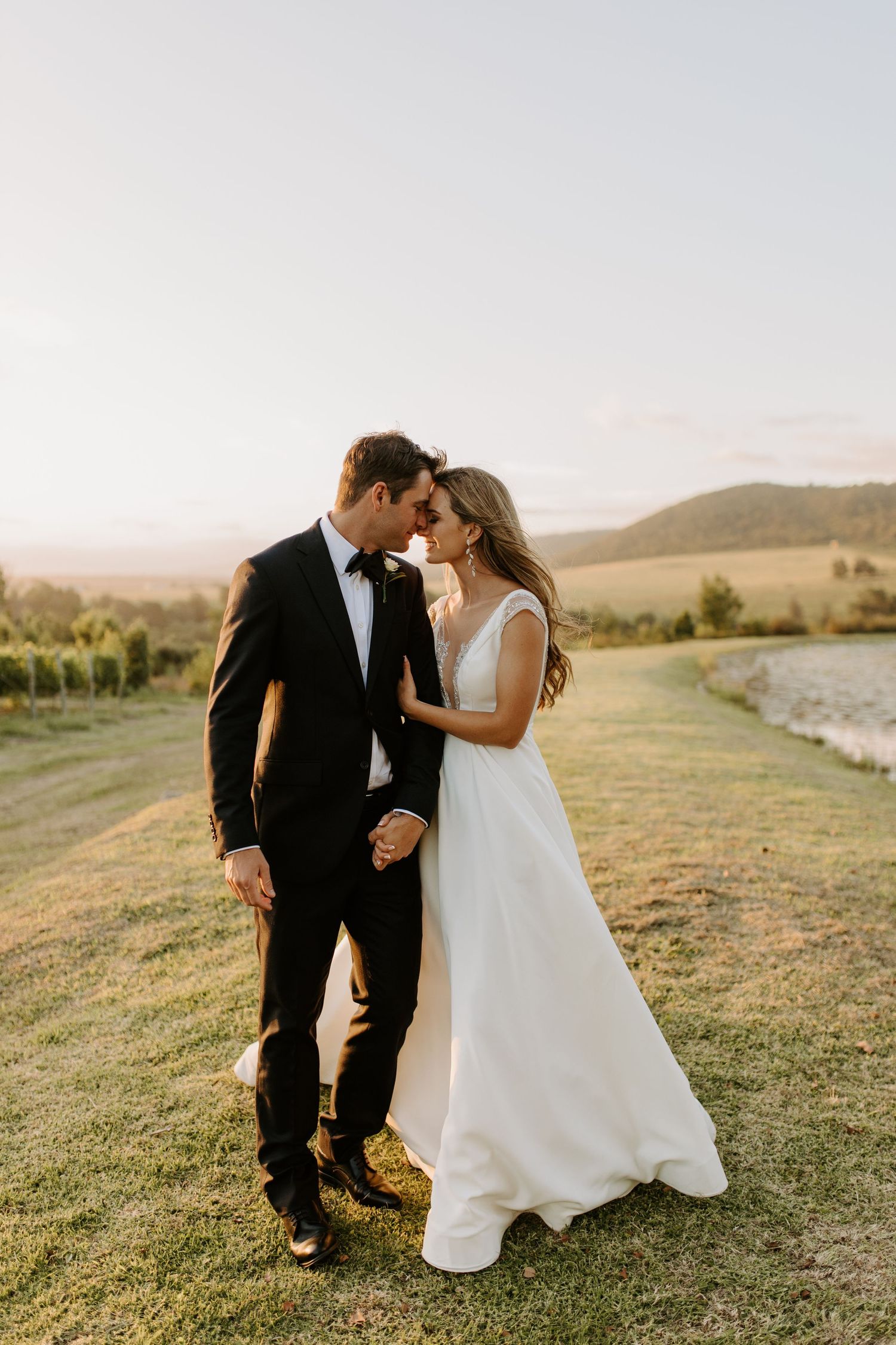 A romantic sunset photo of newlyweds in formal attire embracing on a hilltop overlooking scenic countryside.