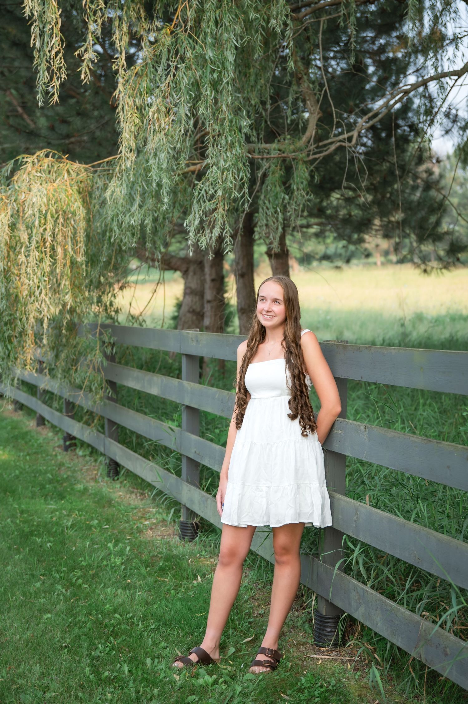 A white lace sundress stands out against a rustic wooden fence and weeping willow trees in a peaceful country setting.