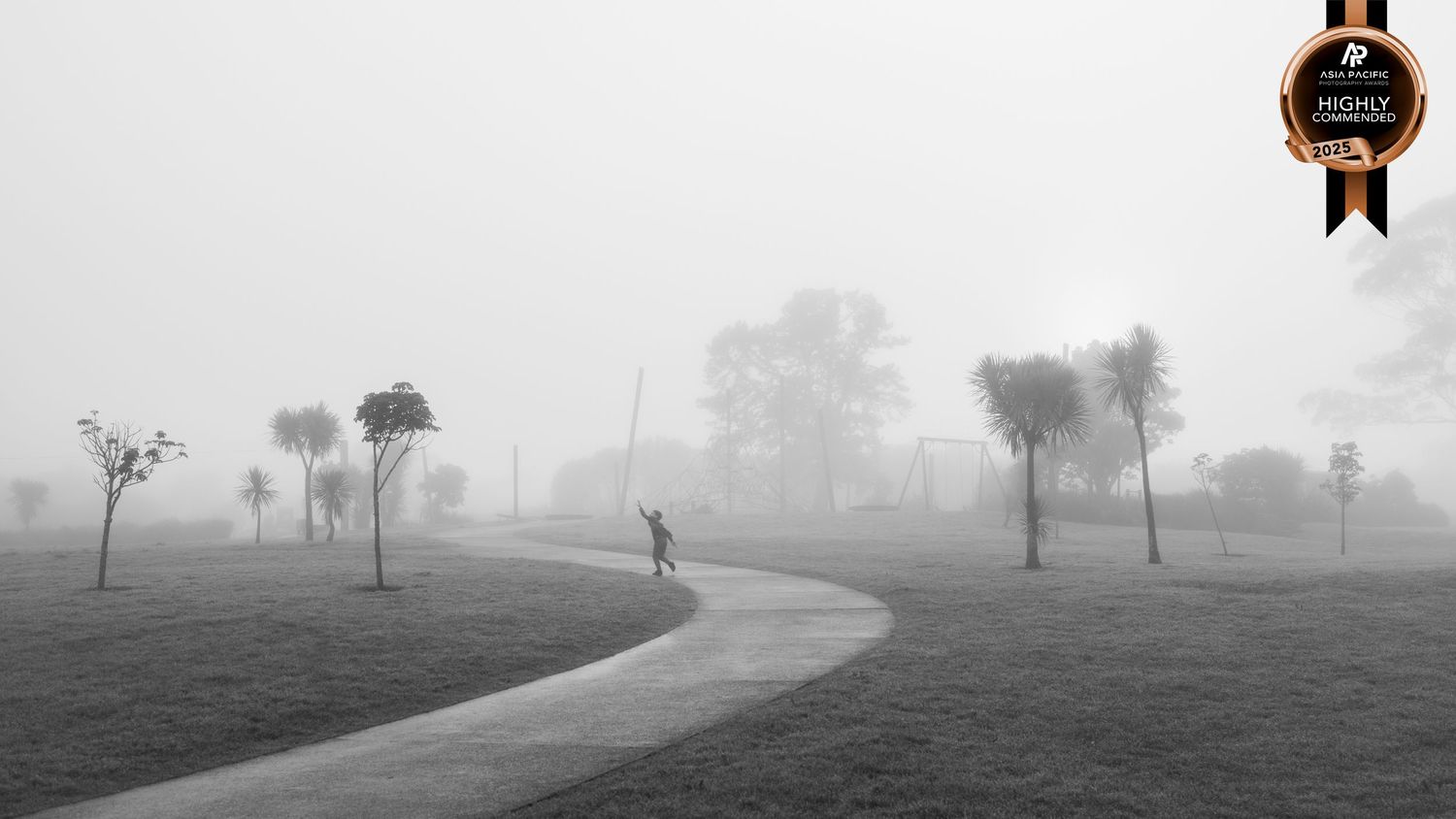 Black and white image of a very misty scene with boy walking on a winding pathway to a playground. He holds his hand up to the mist