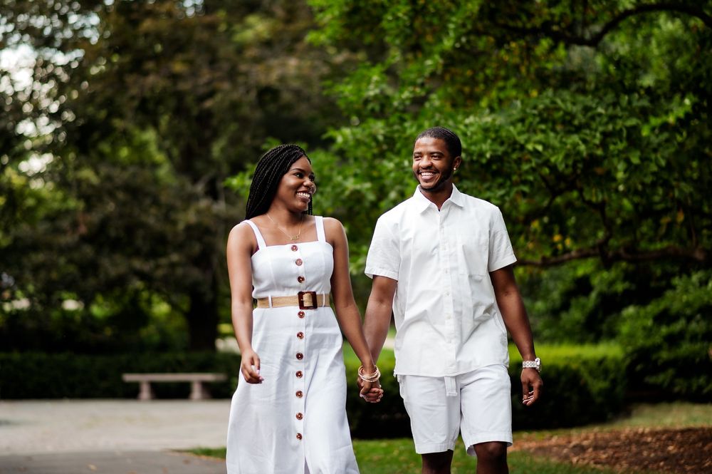 A smiling Black couple walks hand-in-hand through a lush green park, wearing matching white outfits.