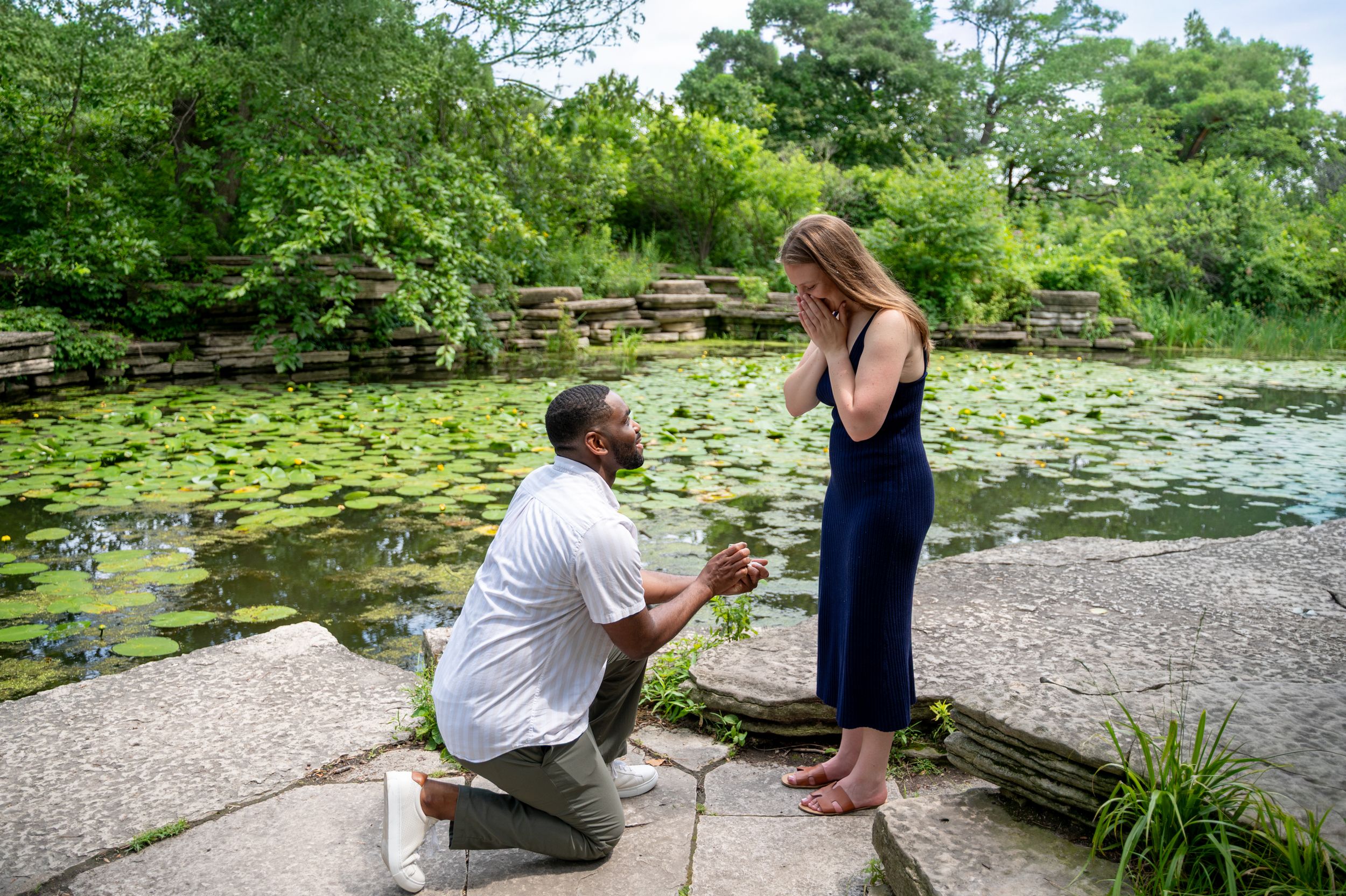 Man proposes to woman on one knee beside a lily pad pond in a lush green garden setting.