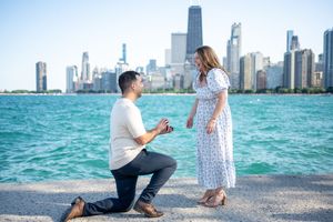 Man proposes on one knee to woman by Lake Michigan with Chicago skyline in background on sunny day.