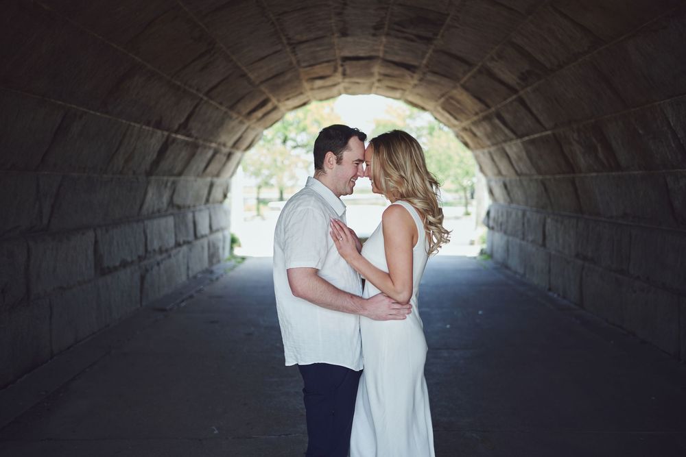 A romantic couple shares an intimate kiss under a stone tunnel arch with soft light filtering in from behind.