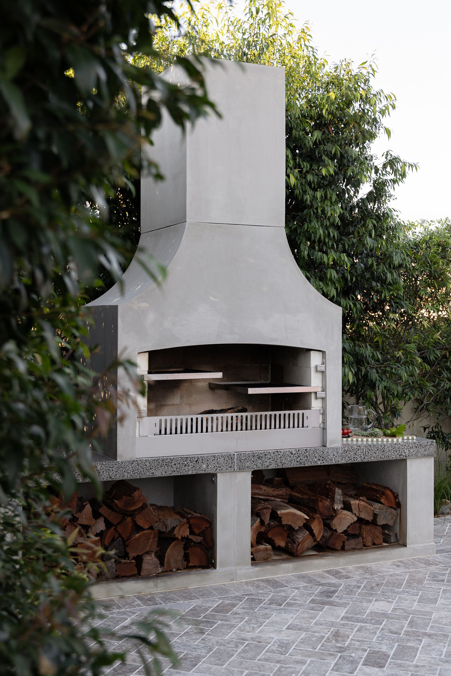 Modern outdoor concrete barbecue grill with firewood storage underneath and stone paver patio in backyard garden setting.