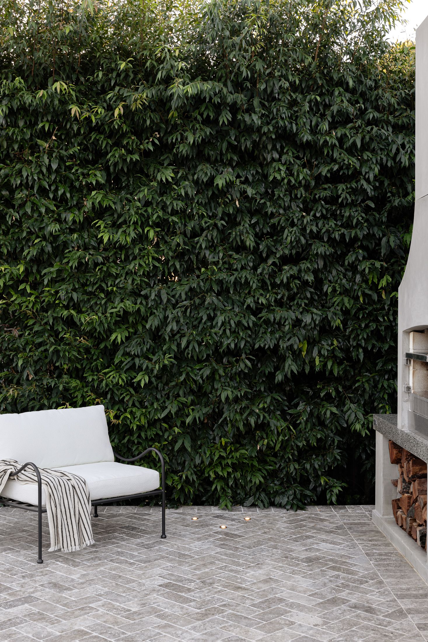 Modern outdoor lounge chair with white cushions sits against a lush green hedge wall in a contemporary patio setting.