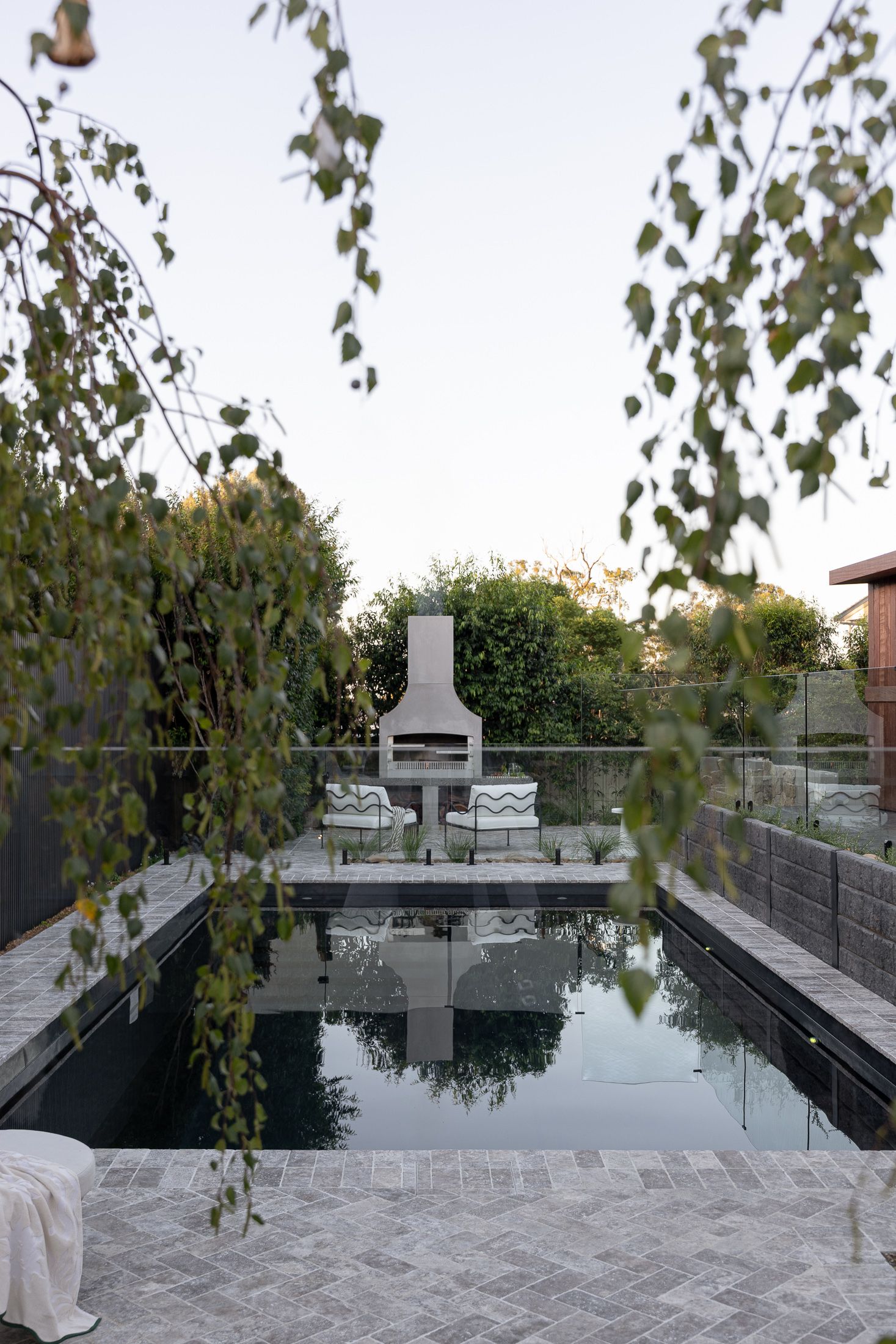 Modern rooftop infinity pool with glass barriers reflects the evening sky while surrounded by lush greenery and city views.