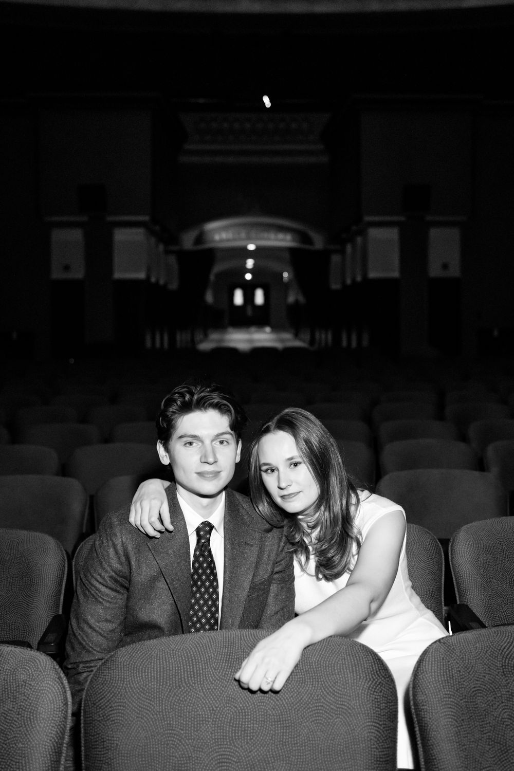 Black and white photo of a young couple sitting in empty theater seats, smiling warmly at the camera.