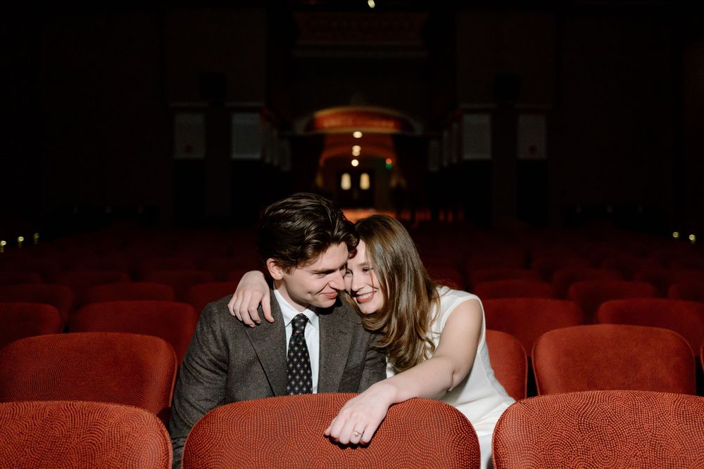Couple sharing a romantic moment in red theater seats, bride in white dress and groom in gray suit.