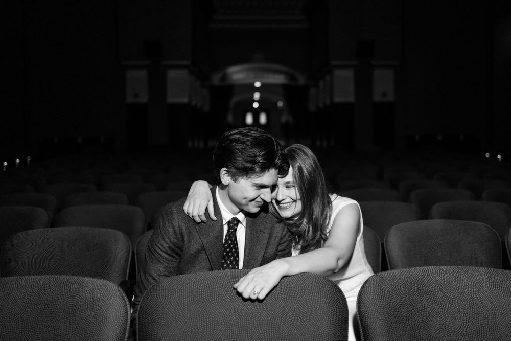 Black and white photo of a romantic couple sitting close together in empty theater seats, sharing an intimate moment.