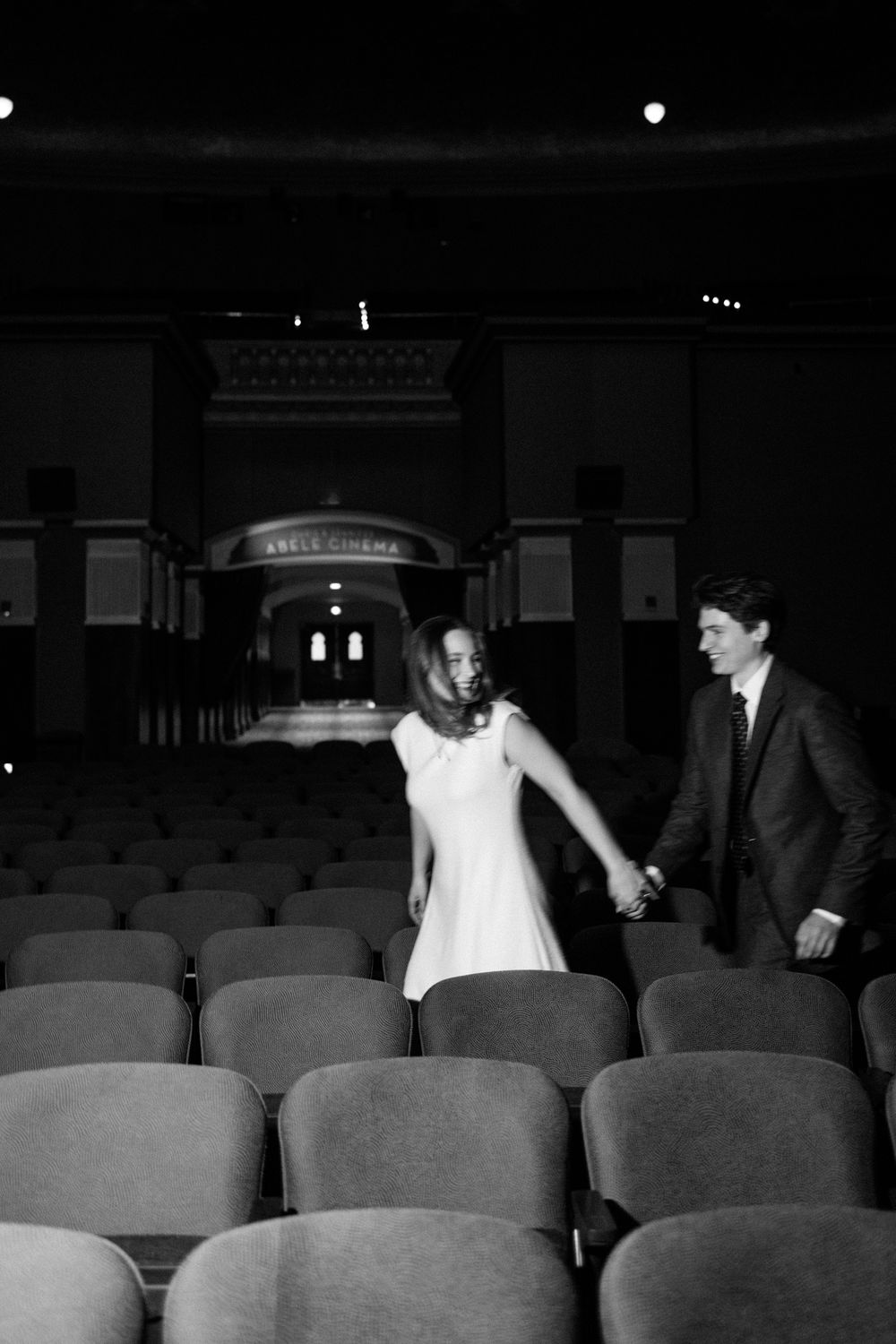 Black and white photo of couple holding hands walking through theater aisle.