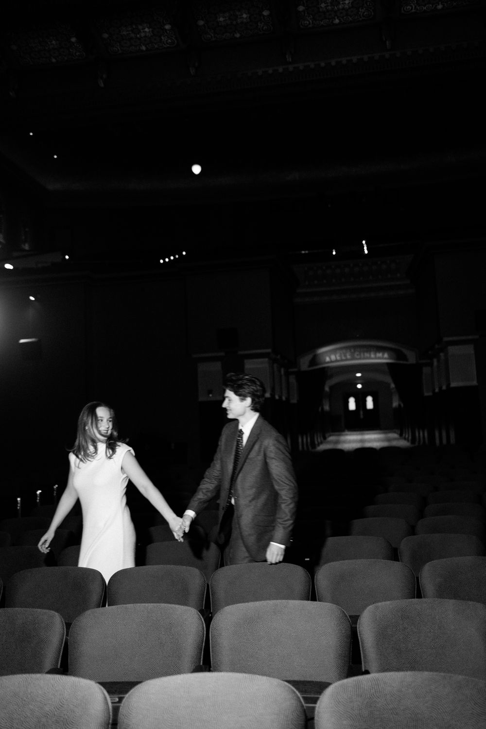 A couple holds hands walking through empty theater rows in an elegant venue, captured in black and white.