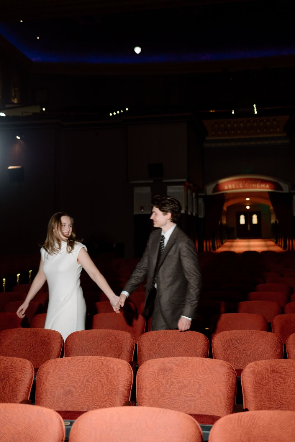 Bride and groom holding hands strolling through rows of red velvet theater seats.
