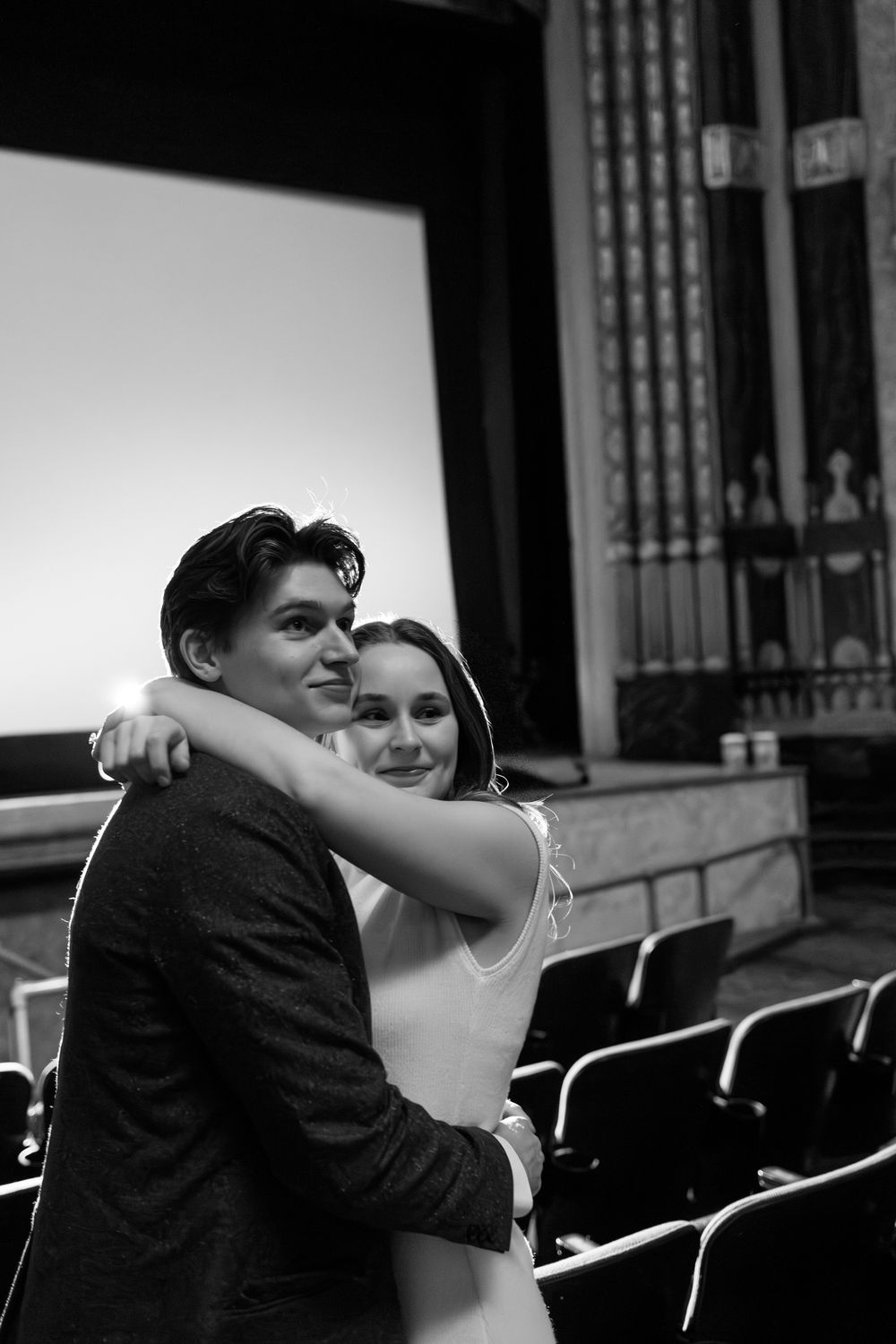 Couple embracing in black and white photo inside elegant theater with ornate decor and blank screen.