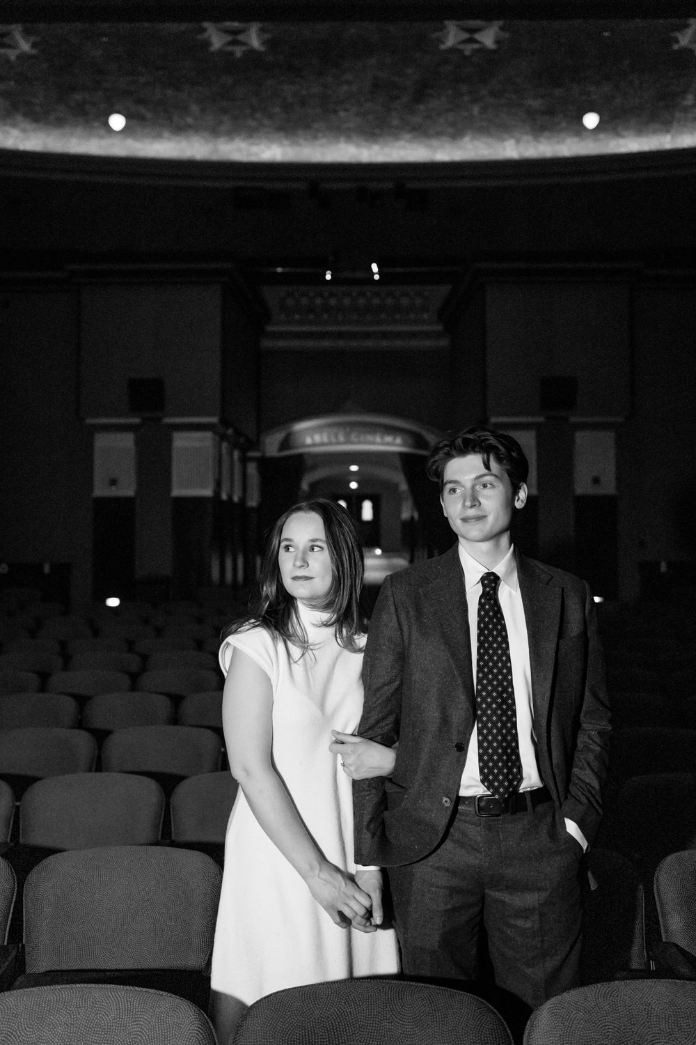 Black and white portrait of elegant couple standing among theater seats in classic cinema.