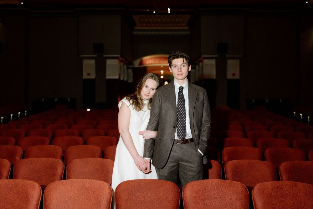 Couple poses among red theater seats, woman in white dress and man in gray suit with tie.