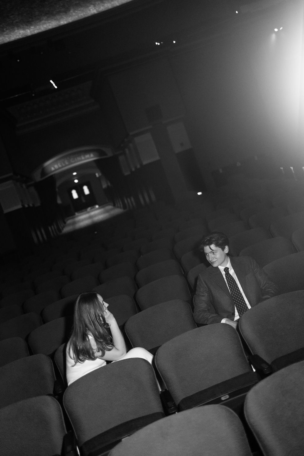 Man and woman sitting apart in empty theater seats sharing an intimate conversation in black and white.