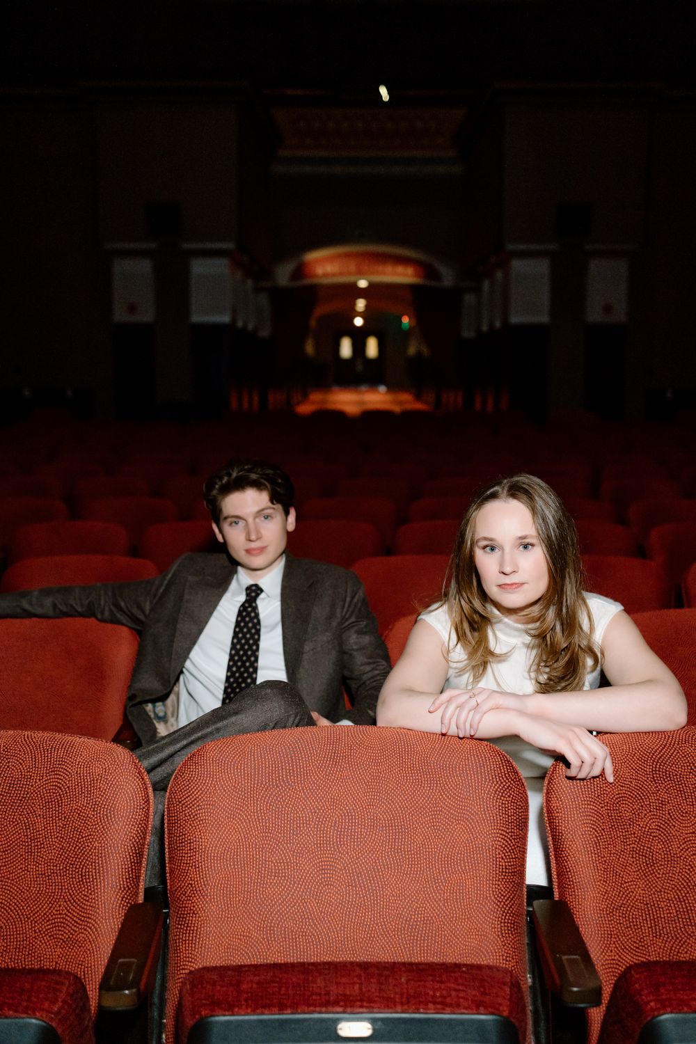 A young man in a suit and a woman in white sit among red theater seats in an empty auditorium.