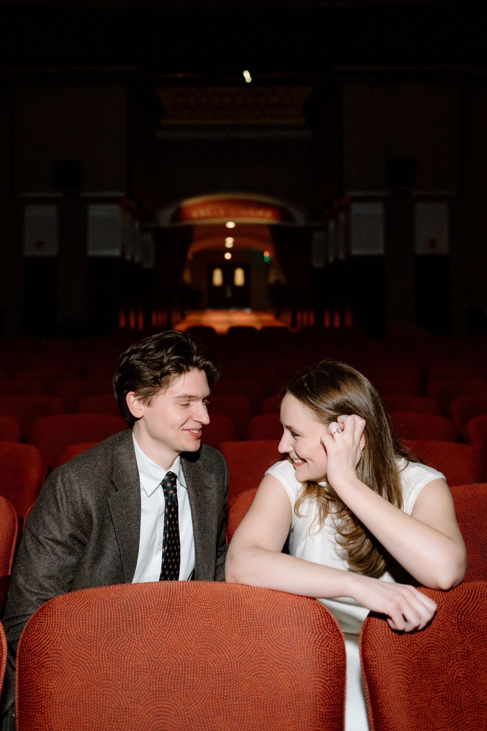 A couple smiles at each other in red theater seats inside an elegant vintage cinema.