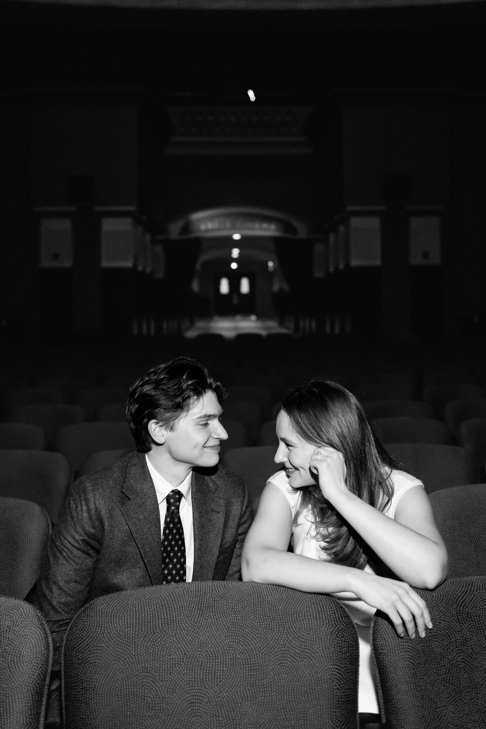 Black and white photo of a couple sharing a romantic moment in vintage theater seats, smiling at each other.