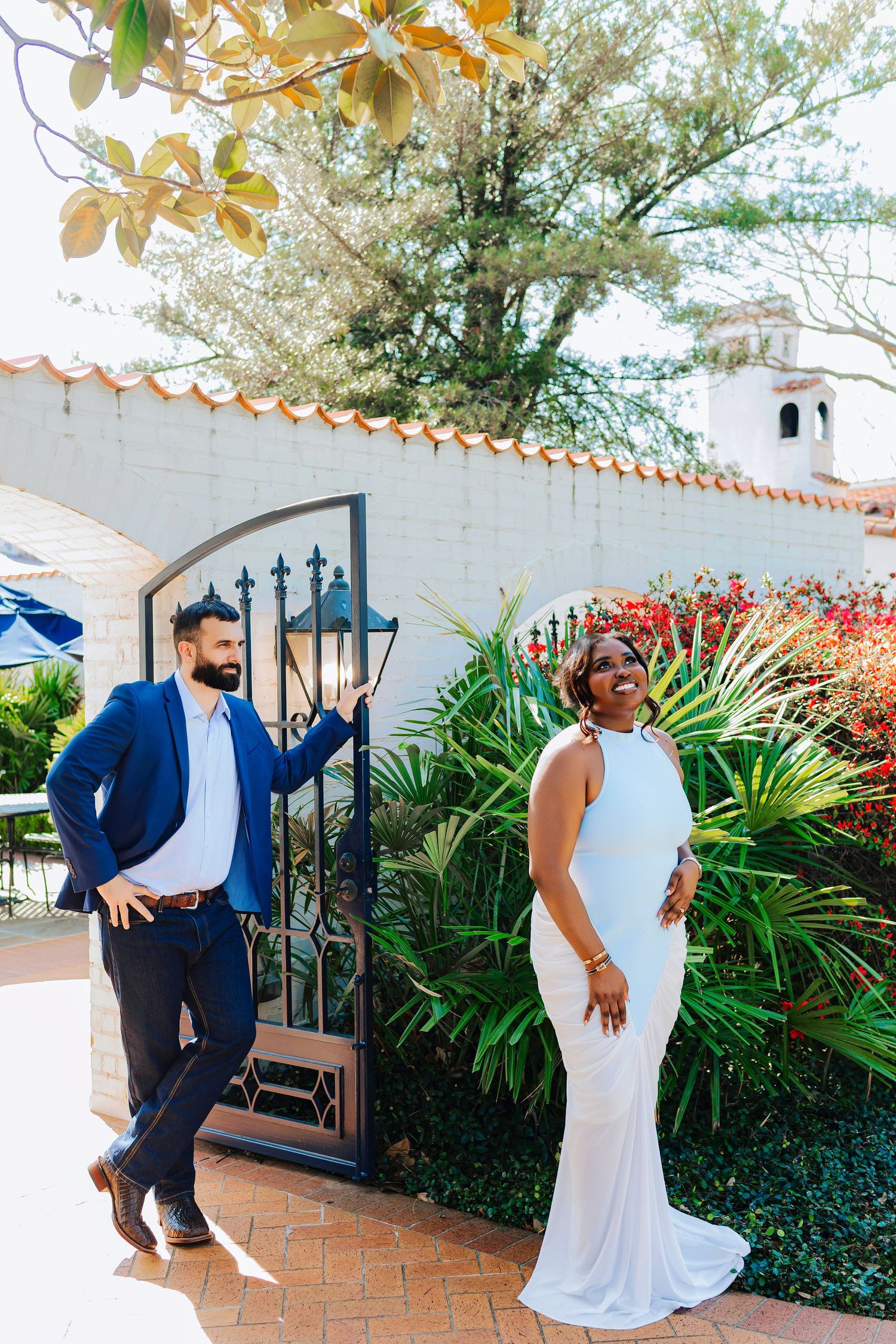 Bride in white gown and groom in blue suit pose by ornate iron gate with lush tropical garden backdrop. at Dallas Arebotum