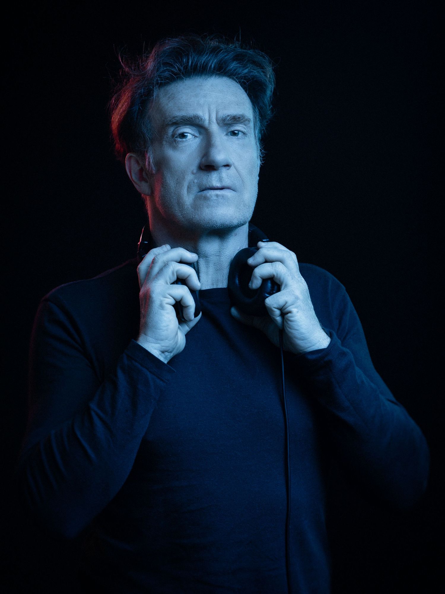 A man in a dark blue sweater holds headphones against his chest under dramatic blue and red lighting.
