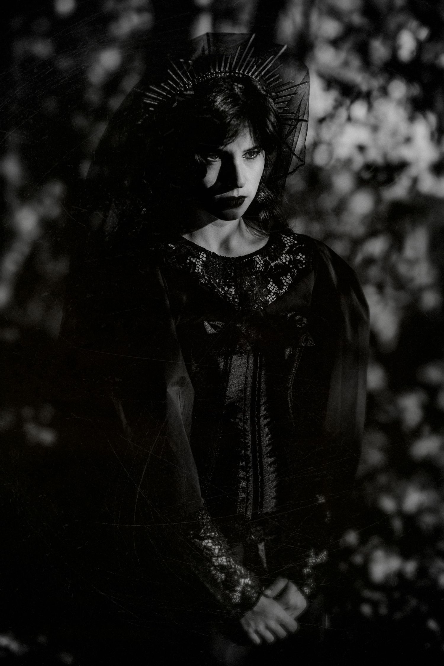 Black and white portrait of a woman in dark gothic attire with a halo crown, surrounded by shadowy trees.