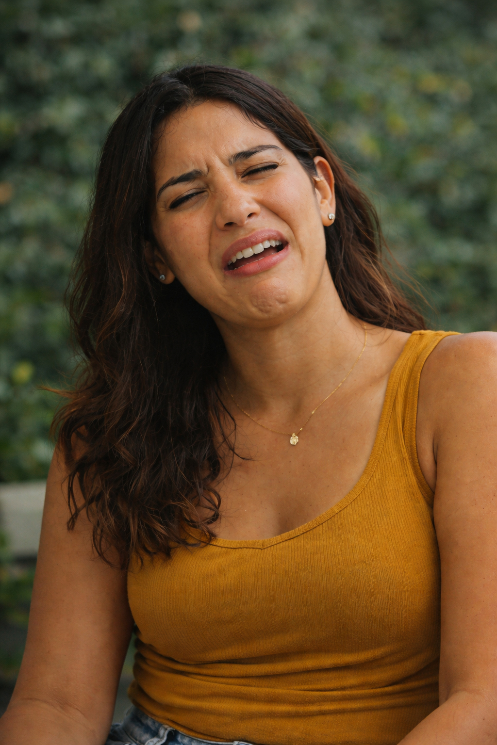 Young woman with dark hair wearing a mustard tank top making a pained expression outdoors.