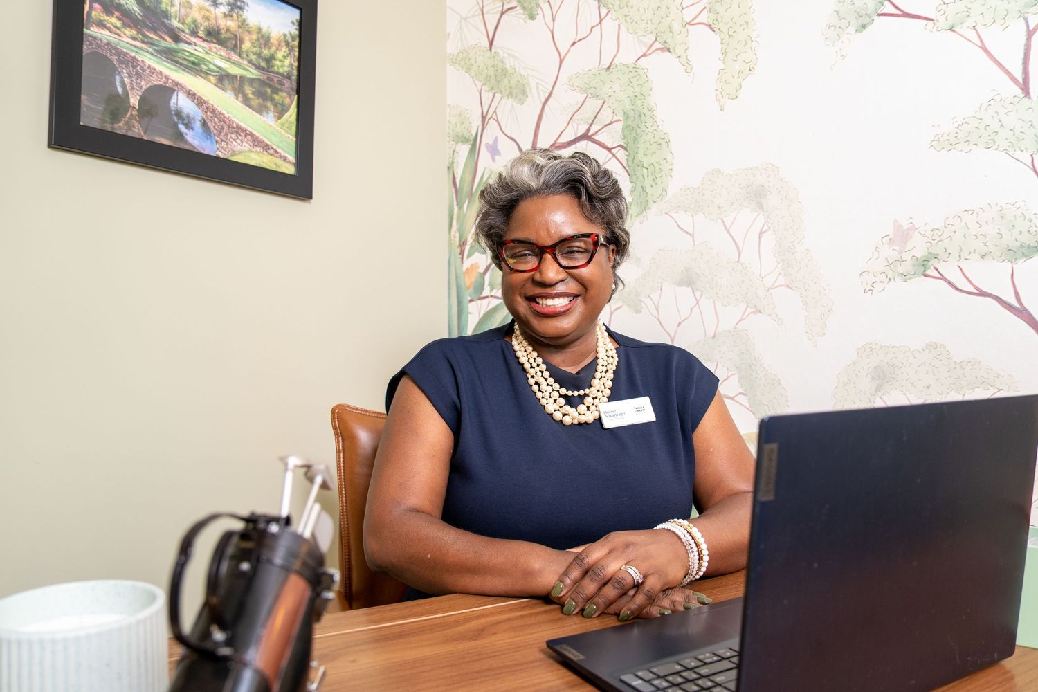 Smiling woman with glasses and a pearl necklace sits at a wooden desk with a laptop in a bright office with tree-patterned wallpaper.