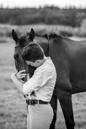 Black and white photo of person embracing horse in field.