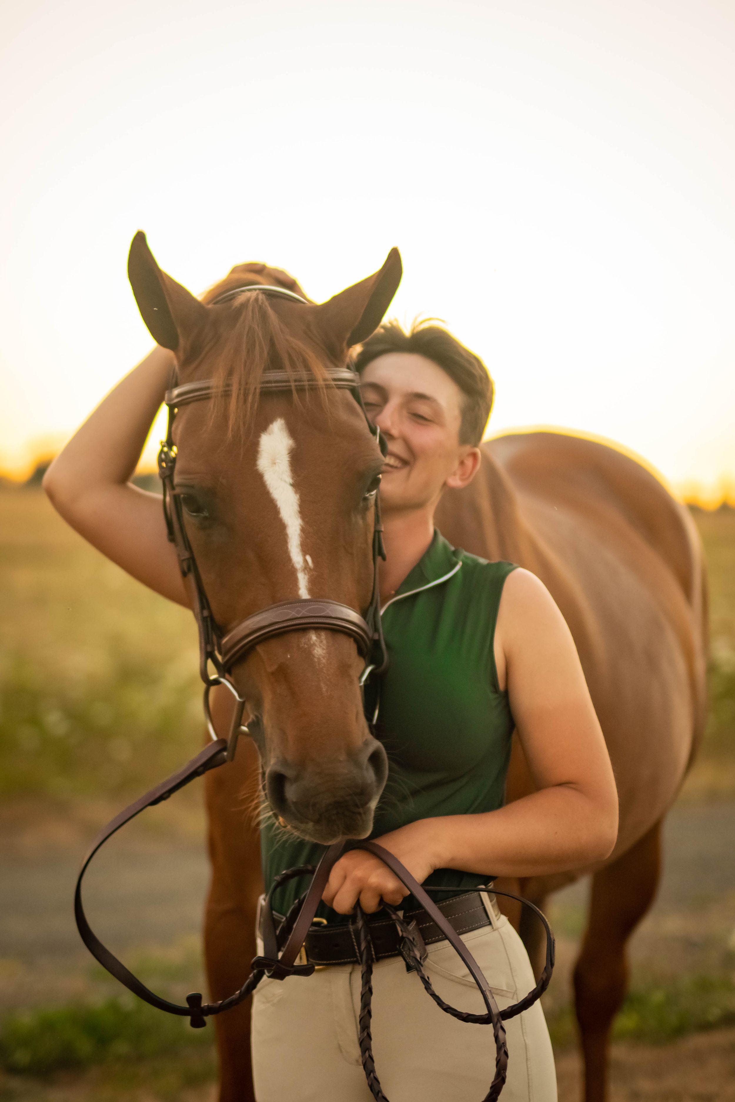 Young equestrian in green vest smiling warmly while petting a chestnut horse at golden sunset.