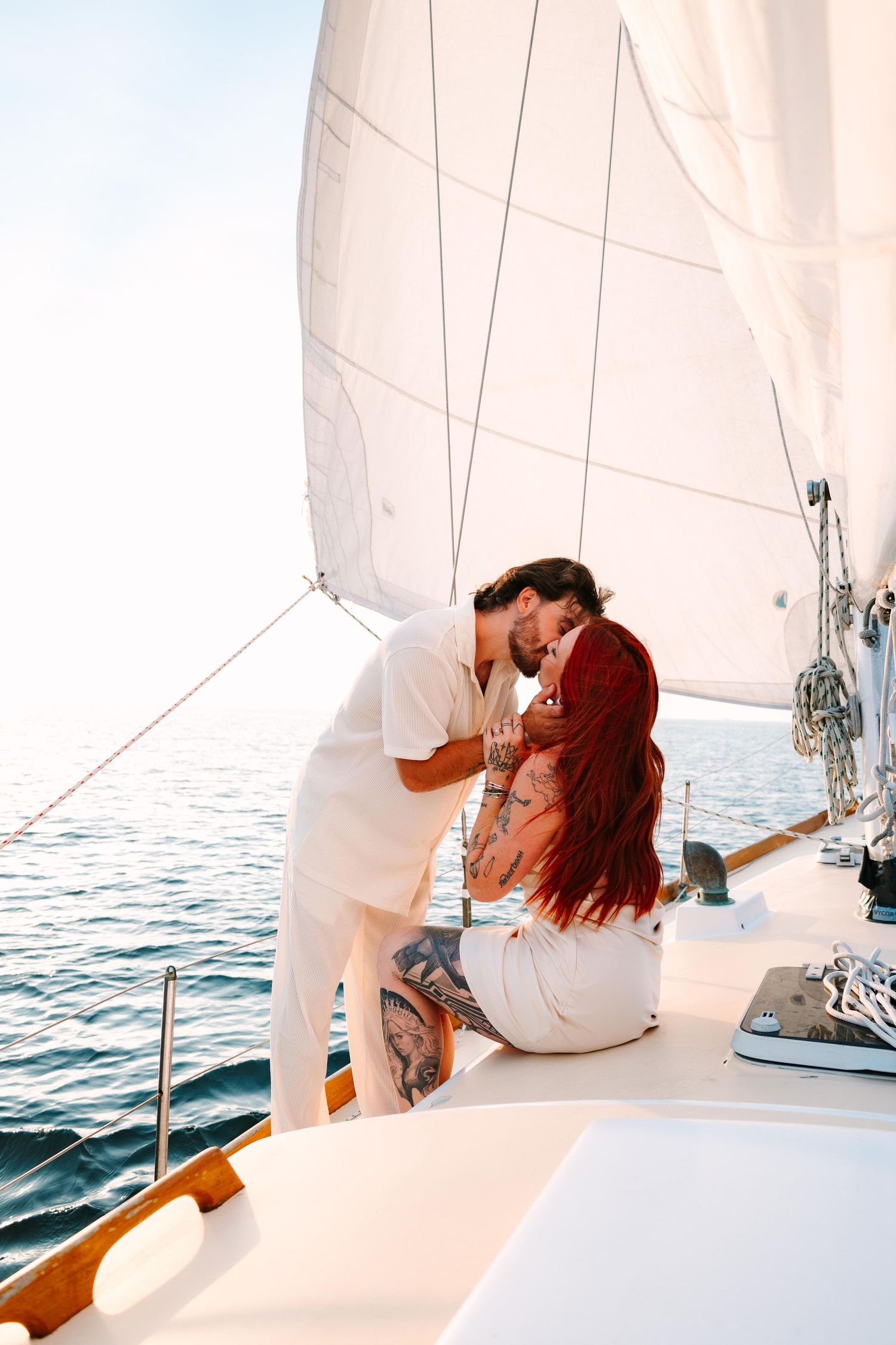 A couple shares romantic moments on a sailboat with white sails and blue ocean waters in the background.