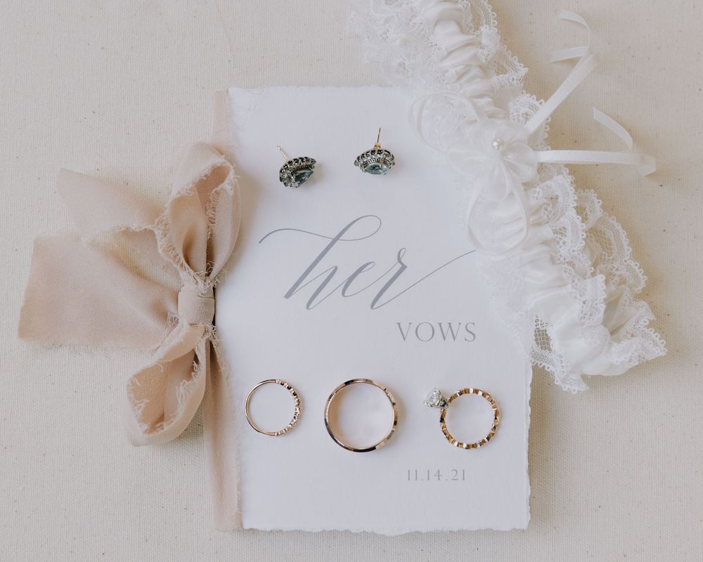 Elegant wedding flat lay featuring vow booklet, gold rings, stud earrings, silk ribbon, and lace garter.