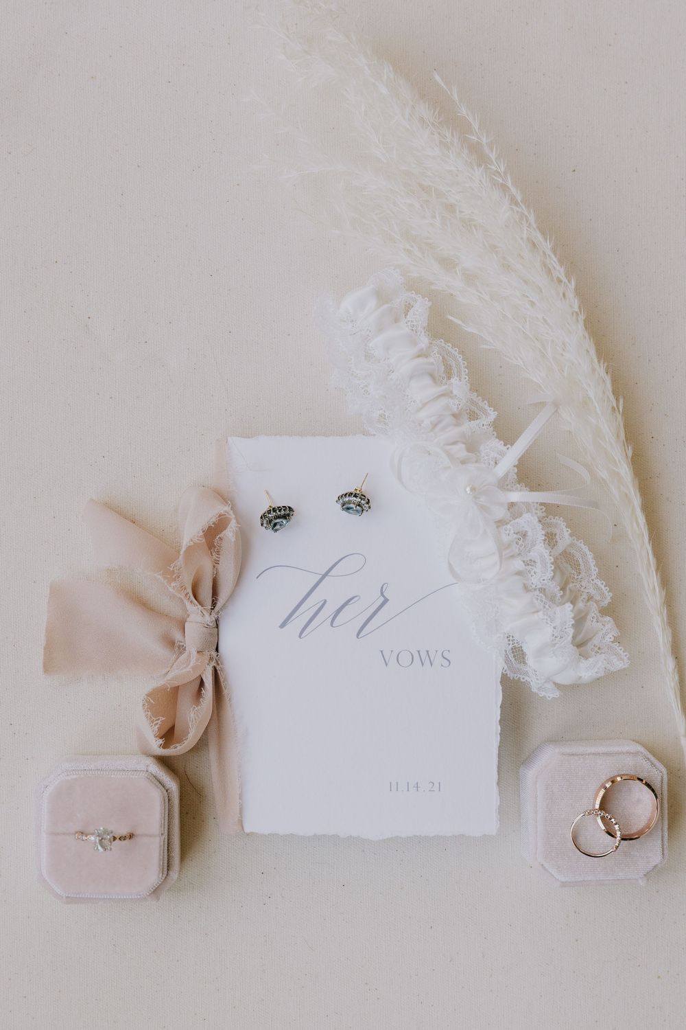 Elegant bridal flat lay with vow booklet, diamond earrings, wedding rings in velvet boxes, lace garter, and pampas grass.