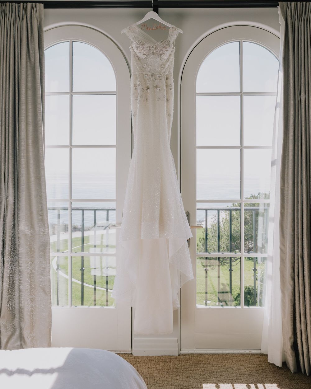Elegant white wedding gown hanging between arched French doors with garden view in bridal suite.