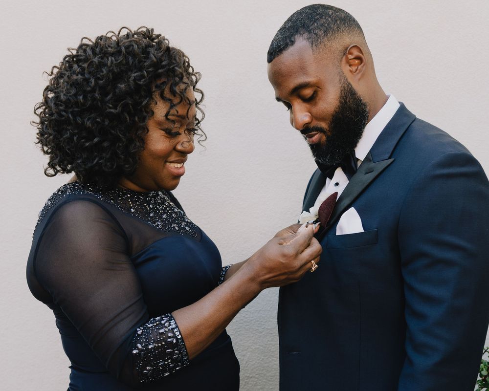 Mother of groom adjusts boutonniere on son's navy suit jacket before wedding ceremony.