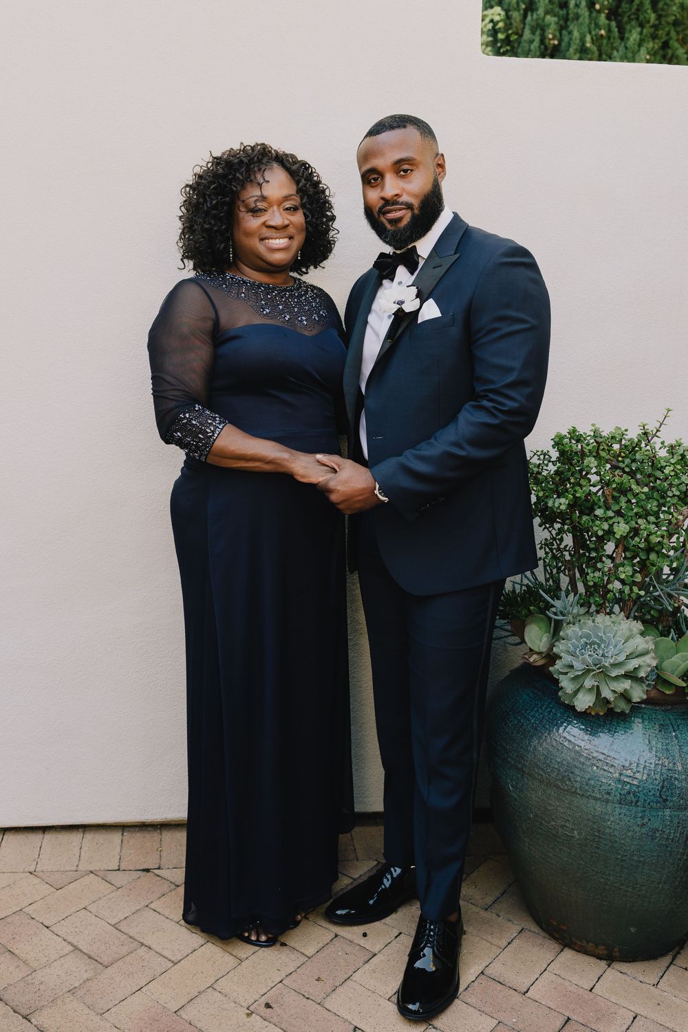 Groom in navy tuxedo poses with woman in elegant black gown at wedding.